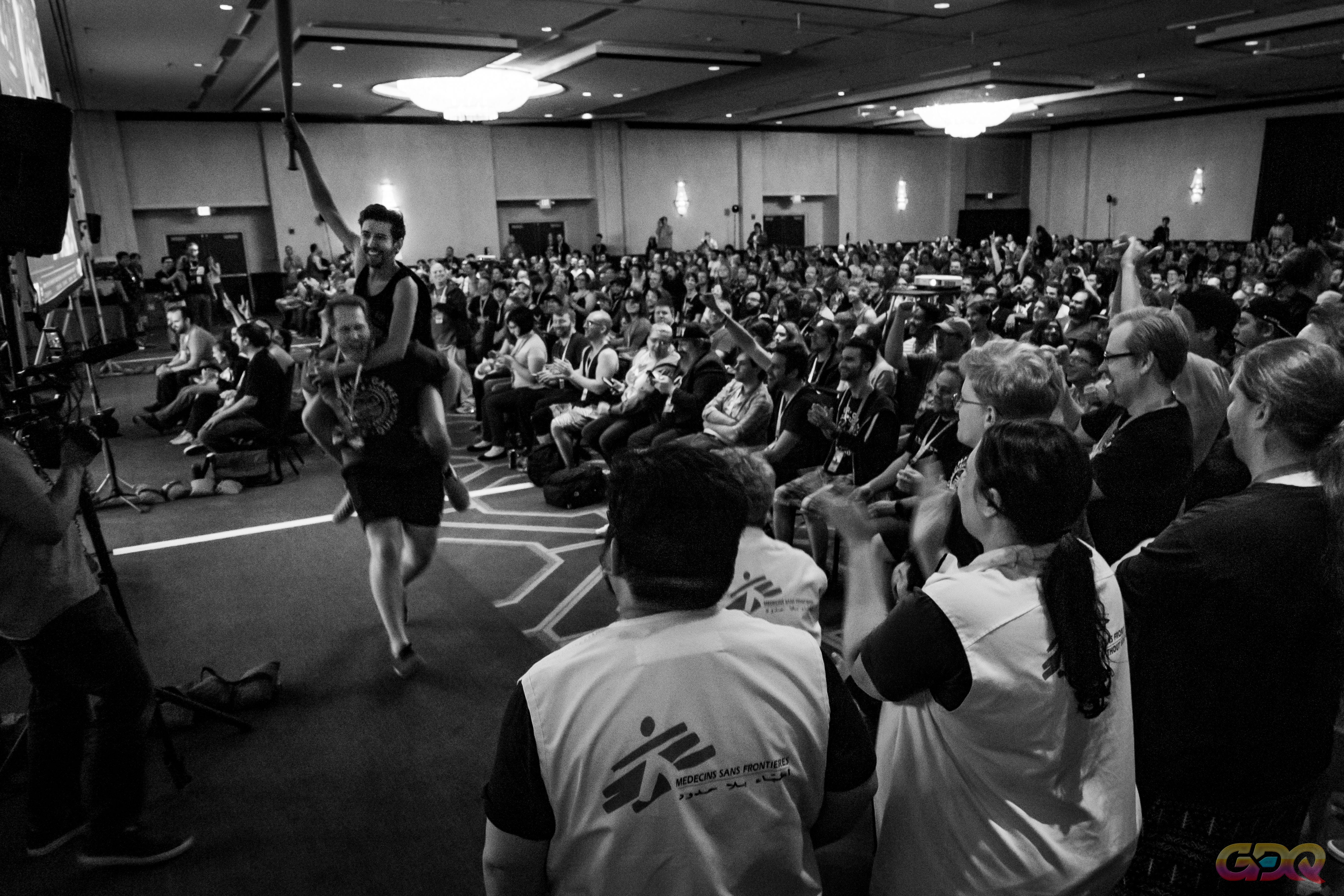 At a live GDQ event, a man is riding on another man piggy-back, holding a baseball bat aloft in the air, both are cheering in front of a large crowd. In the foreground, volunteers from Doctors Without Borders are clapping.